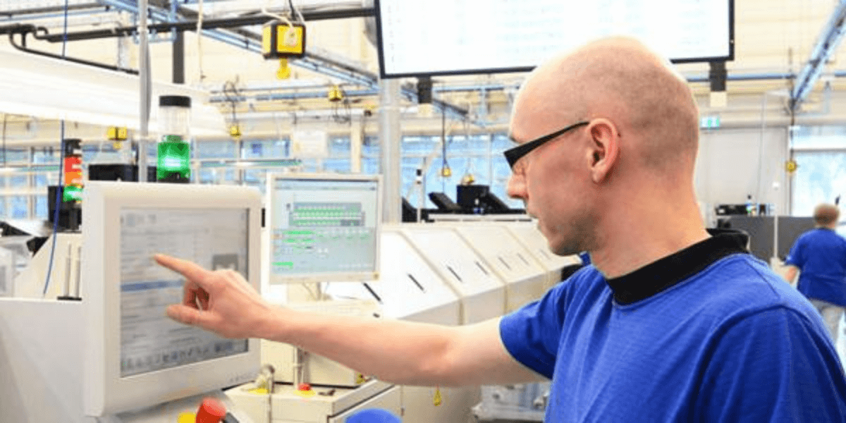 A bald male employee pointing to a computer screen inside of a factory.