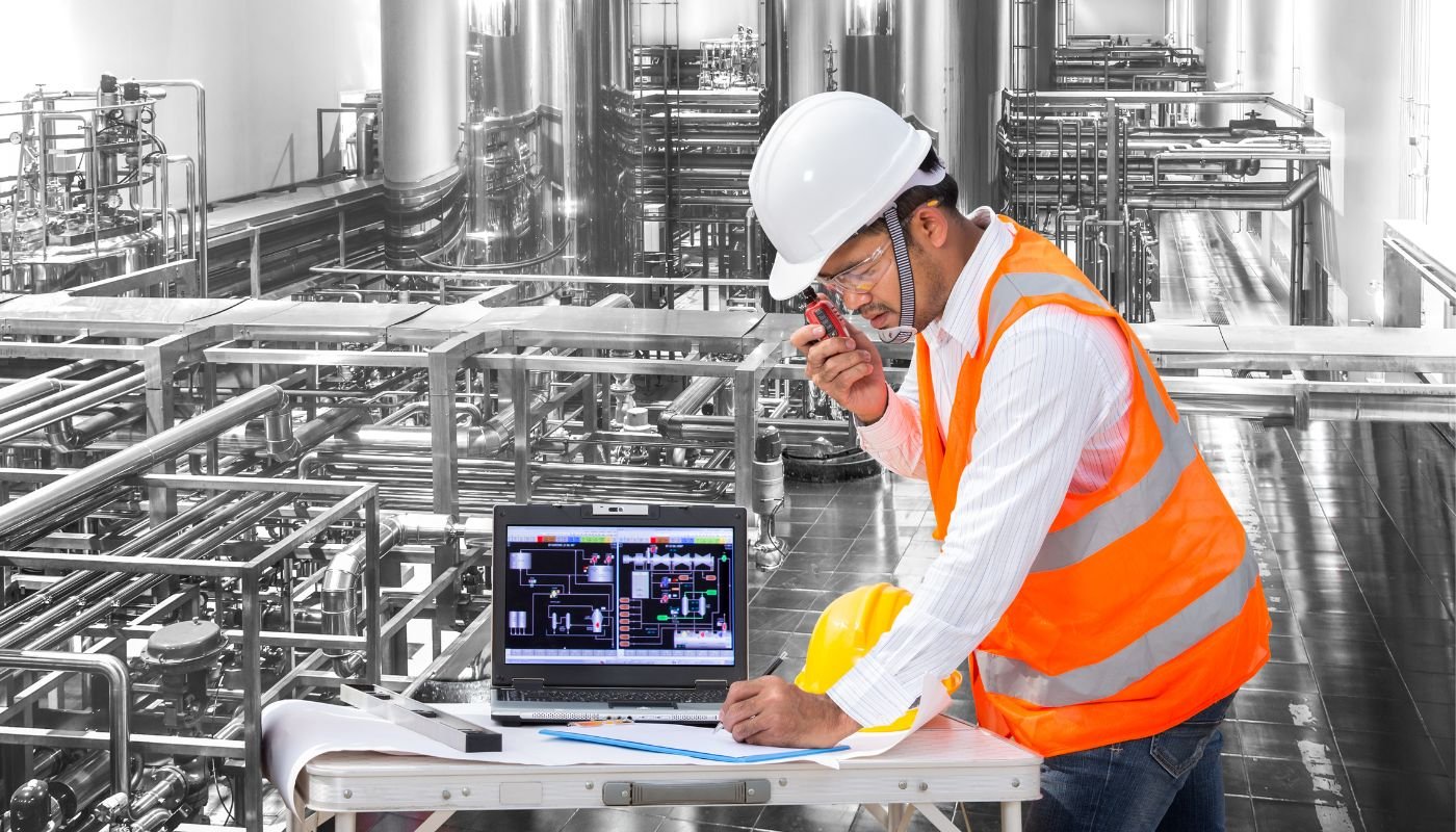 An industrial worker with a white hard hat and an orange vest checking data on a computer in a factory.