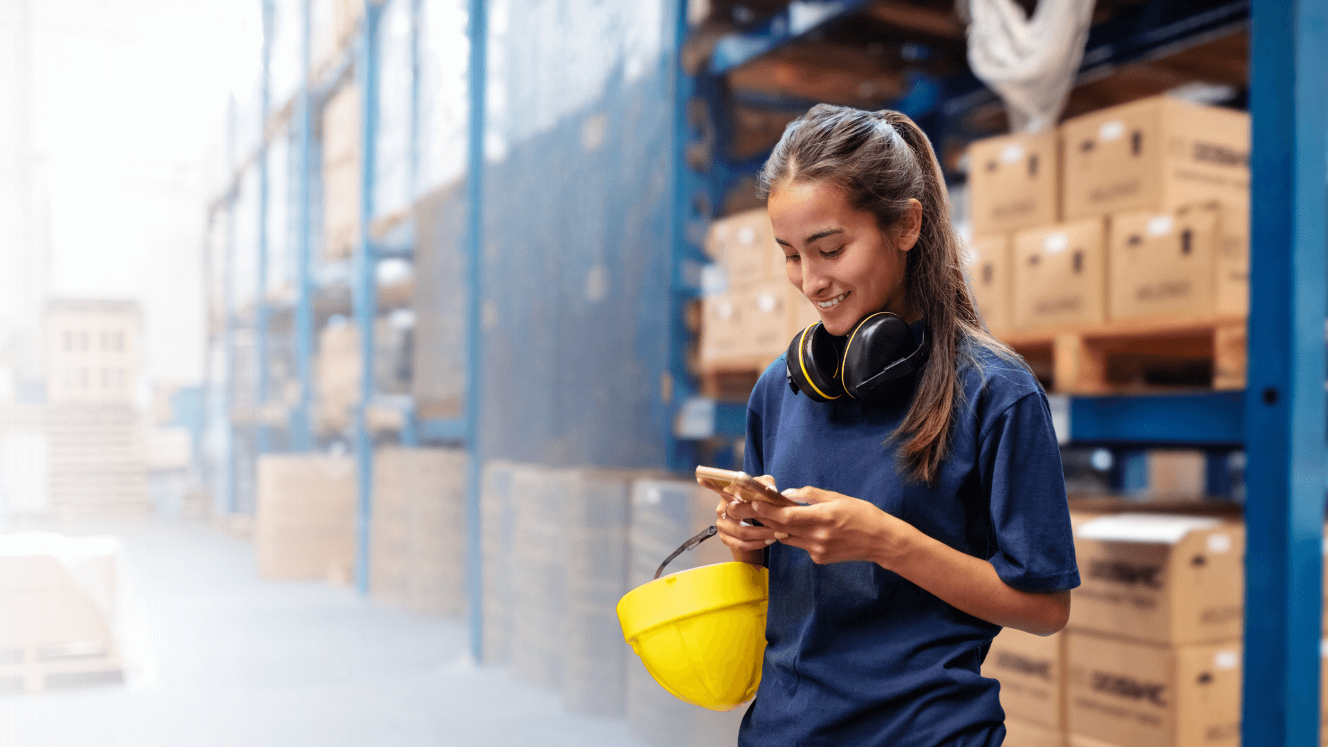 An industrial worker using a cell phone in a warehouse wearing a blue shirt while holding a yellow hard hat. (3) (1)