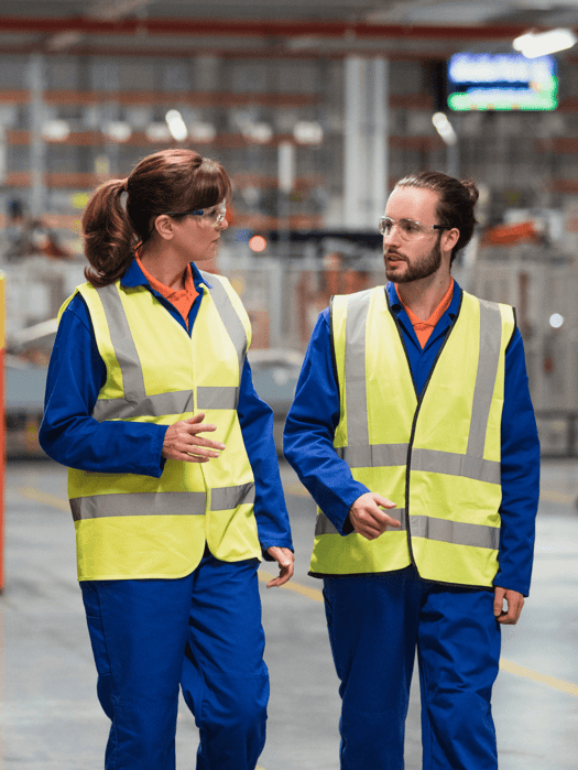 two workers in a factory warehouse wearing blue coveralls and yellow vest talking.