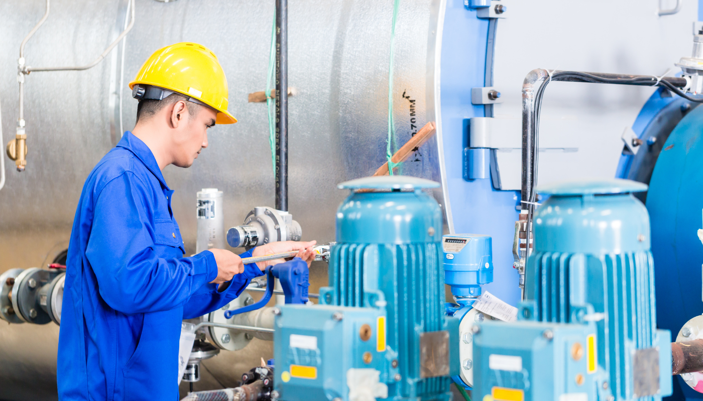 A worker in blue coveralls and a yellow hard hat doing maintenance work on industrial equipment.