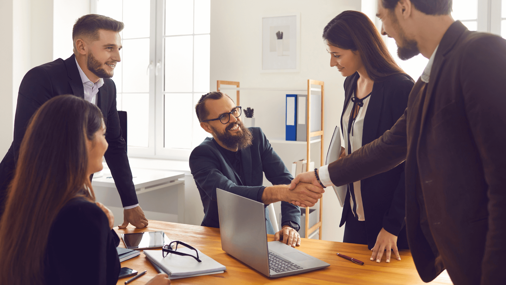 A group of IT business partners shaking hands at a table, making a deal with a computer in front of them on a desk. (1)